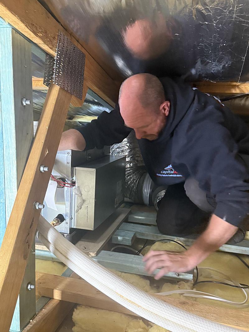 Capital Air technician installing ducted heating and cooling equipment inside a roof space.