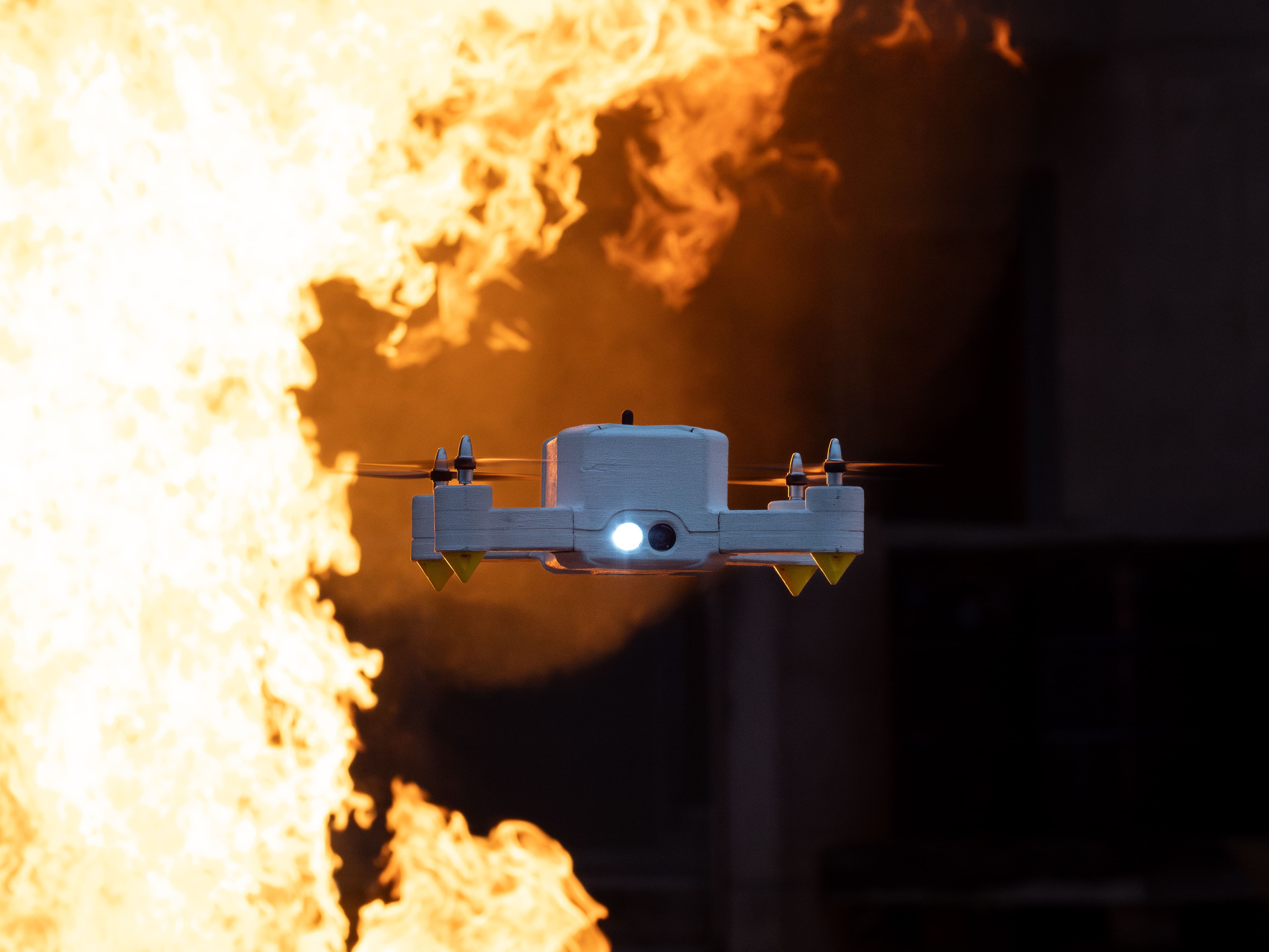 Small white drone with spinning propellers flying in front of large flames.