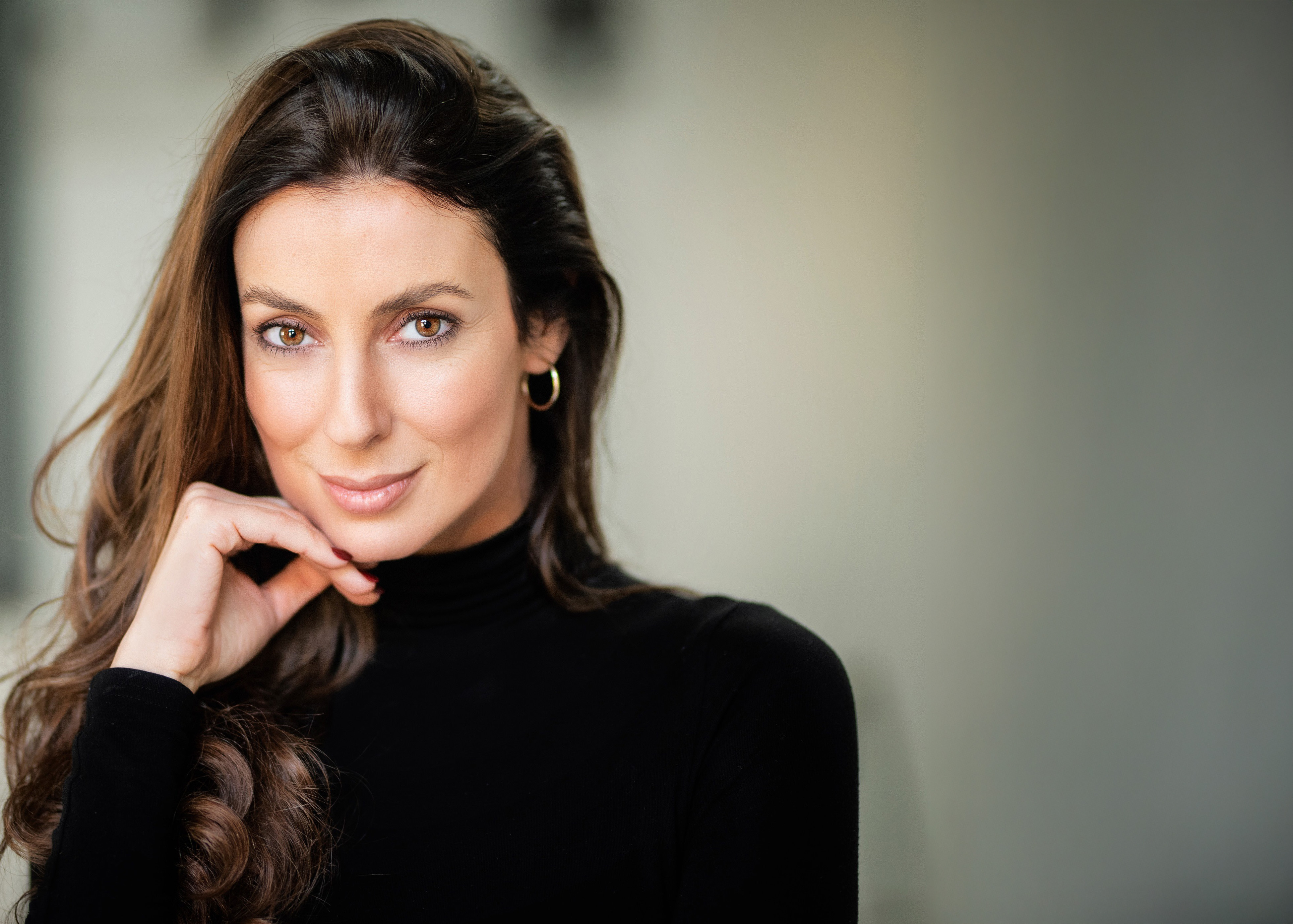 Smiling woman with long brown hair and gold hoop earrings wearing a black turtleneck, resting her chin on her hand.