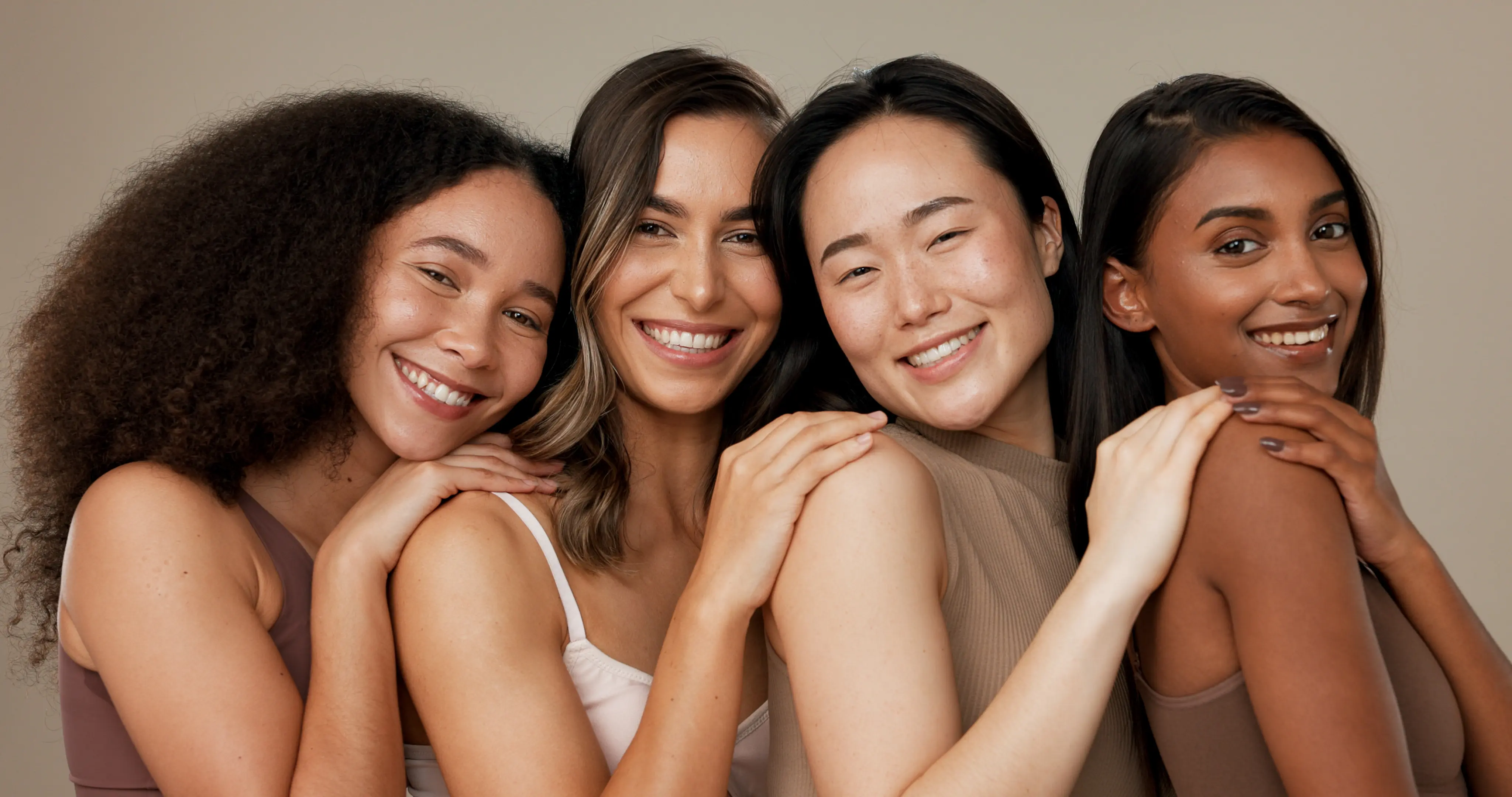 Smiling diverse women standing close together with hands on each other's shoulders against a beige background.