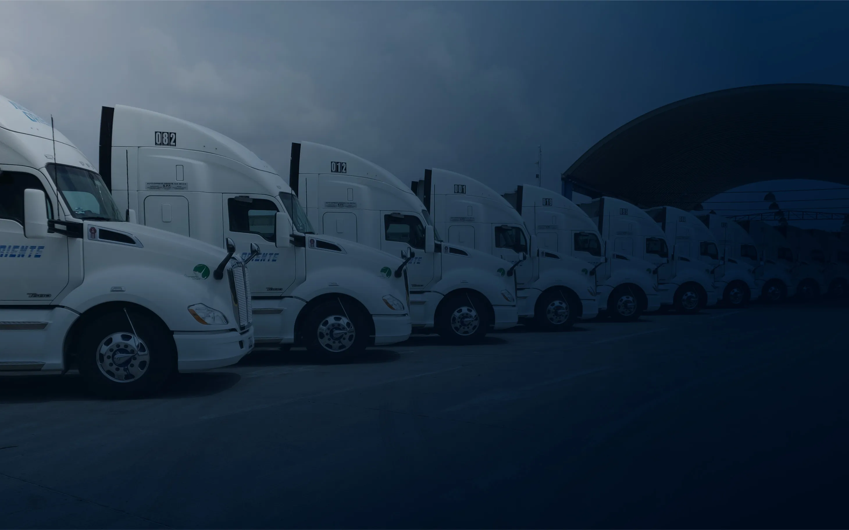A row of white semi-trailer trucks parked under a large covered structure at a truck depot.