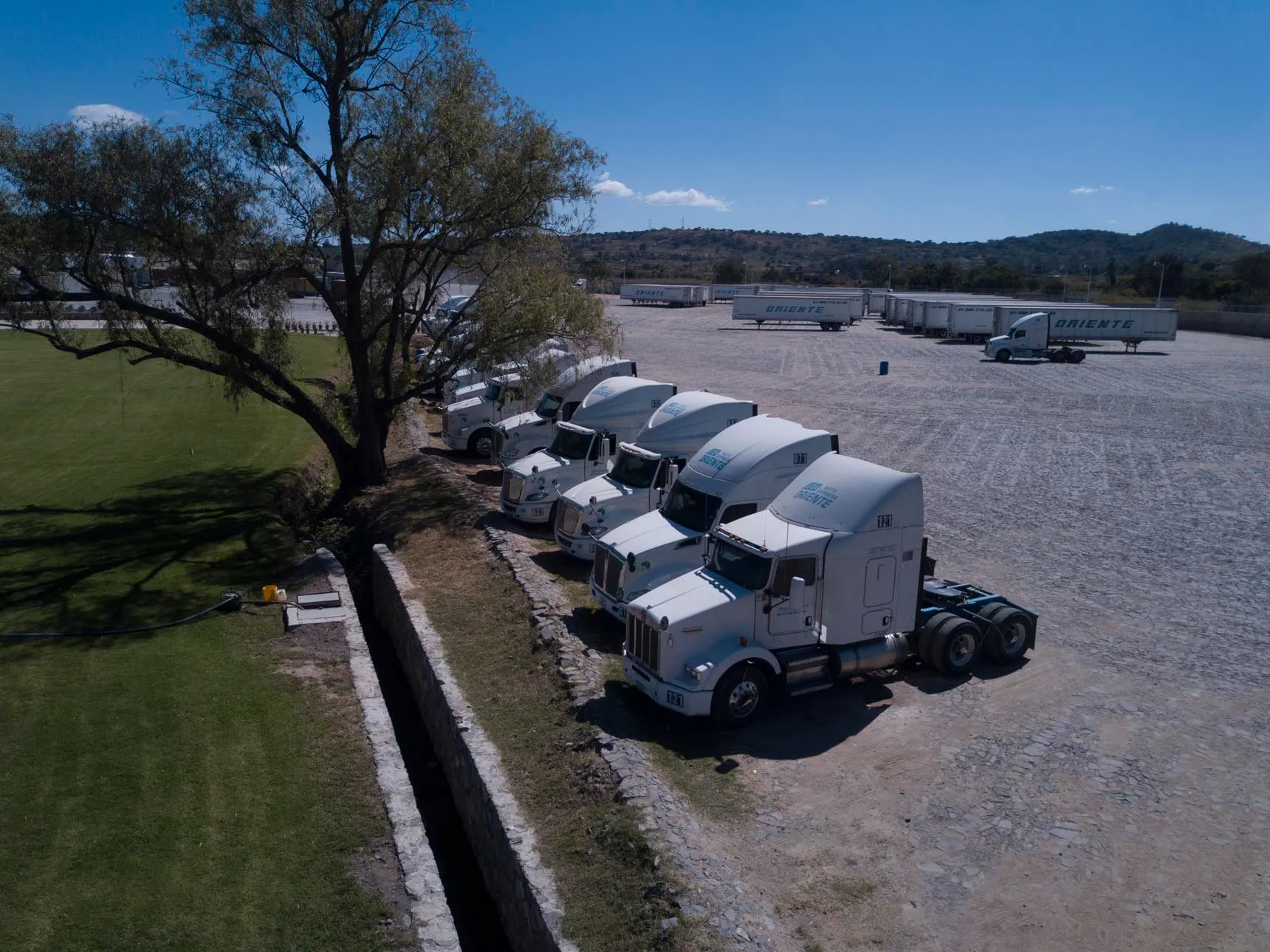 A woman in a wetsuit holding a surfboard on a sandy beach, with surf equipment in the background and tents set up nearby under a clear blue sky.