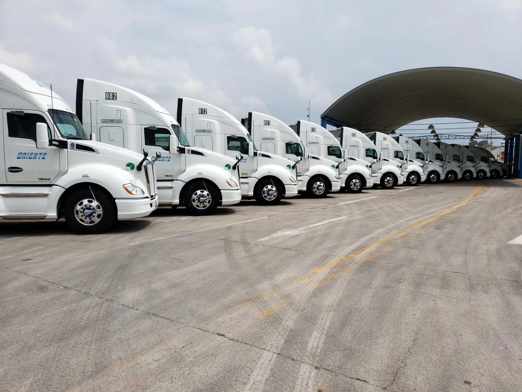 Row of white semi-trucks parked under a large curved shelter in a truck yard.