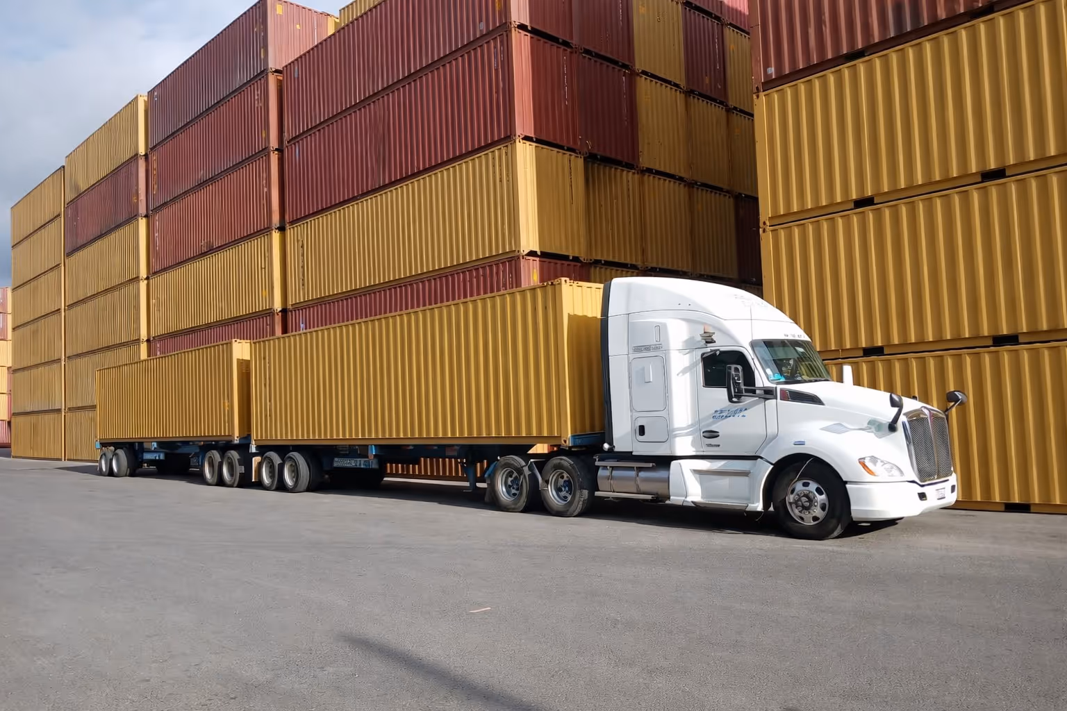 White semi-truck with two large yellow shipping containers parked in front of stacked red and yellow shipping containers.