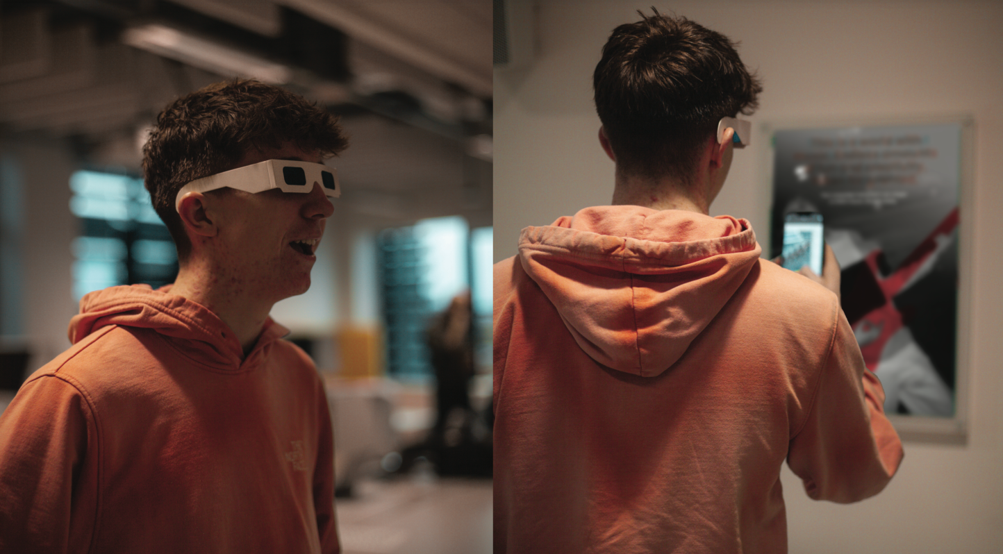 Young man in a peach hoodie wearing white rectangular glasses, shown from side and back views, using a smartphone to scan a poster on a wall.