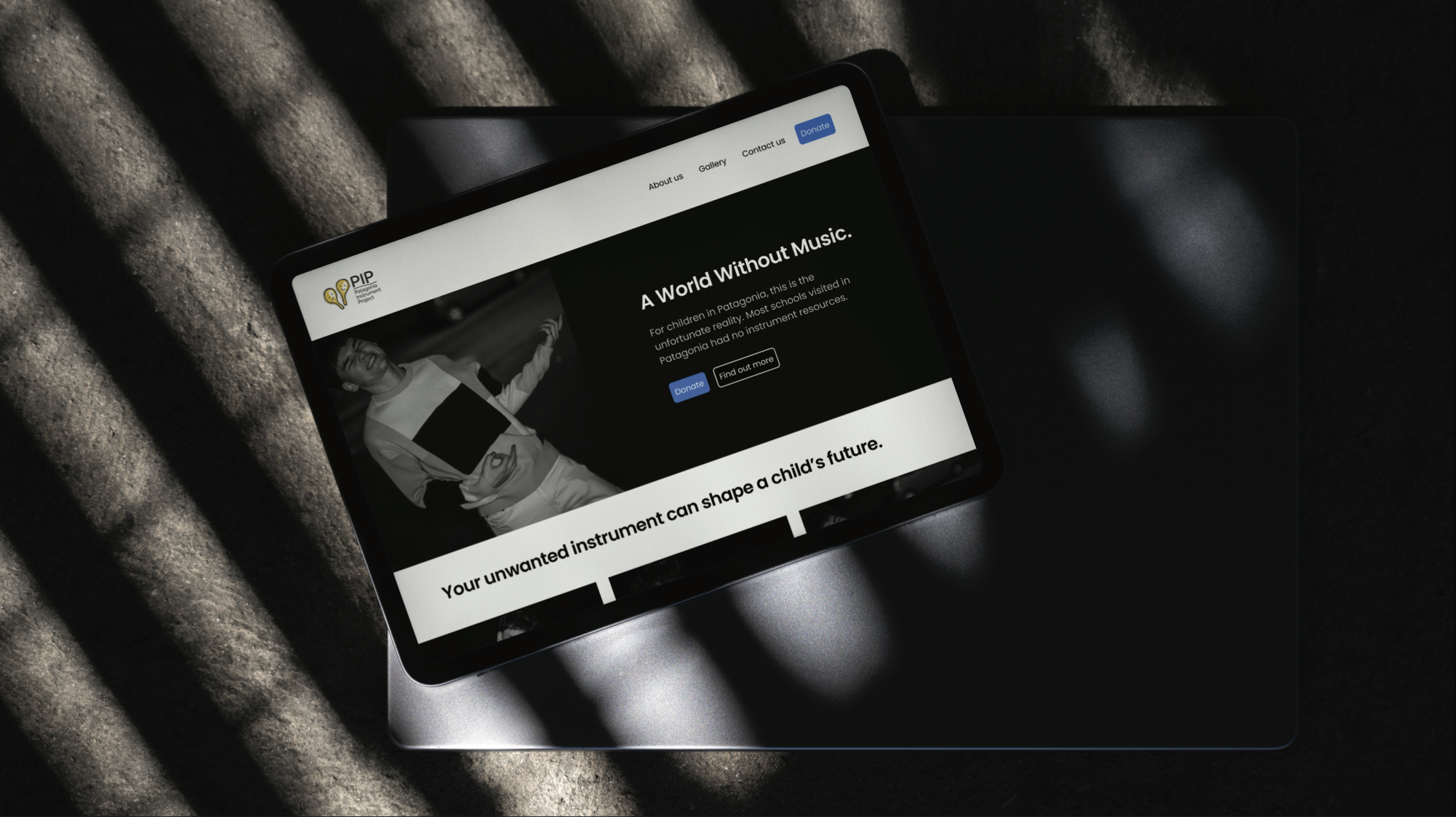 Tablet displaying a website titled 'A World Without Music' focused on donating musical instruments for children in Patagonia, placed on a laptop with shadow stripes across.
