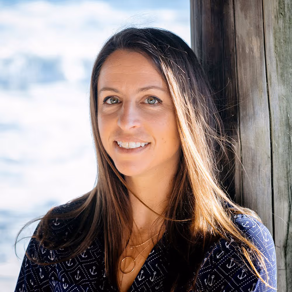 Smiling woman with long brown hair standing beside wooden posts with water in the background.