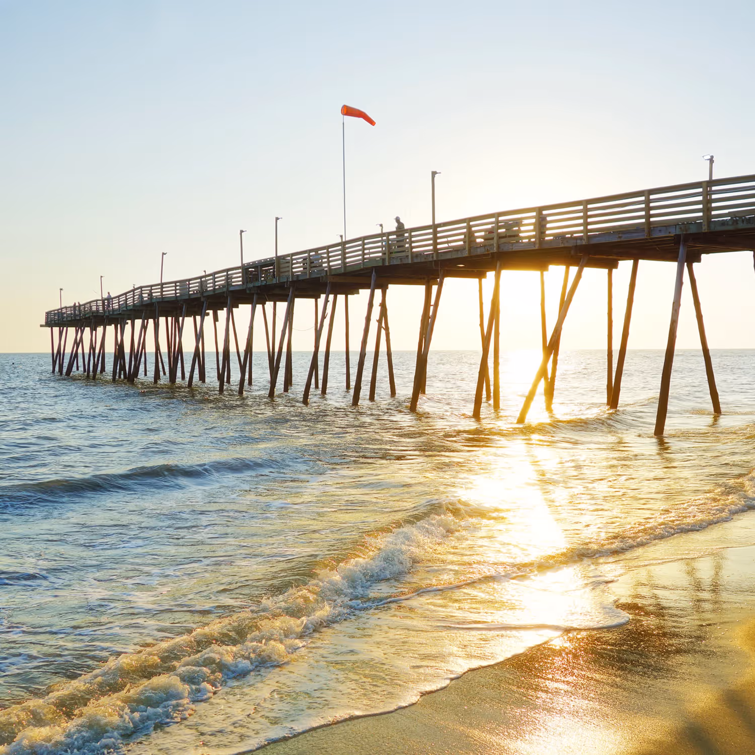 Wooden pier extending over ocean water with a setting sun reflecting on the waves and sandy beach.