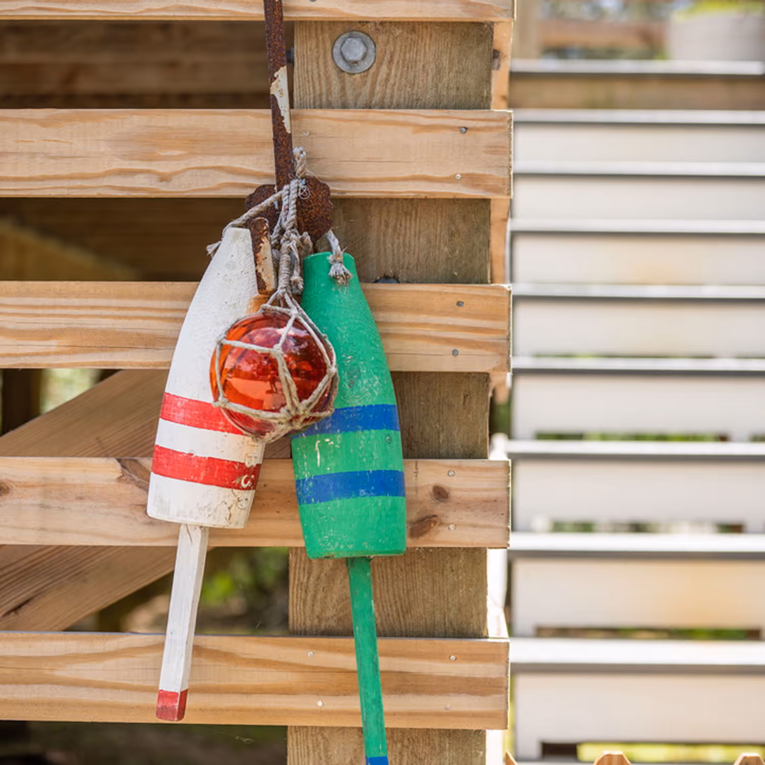 Two wooden fishing buoys, one white with red stripes and one green with blue stripes, hanging on a wooden structure with a red glass fishing float wrapped in rope.
