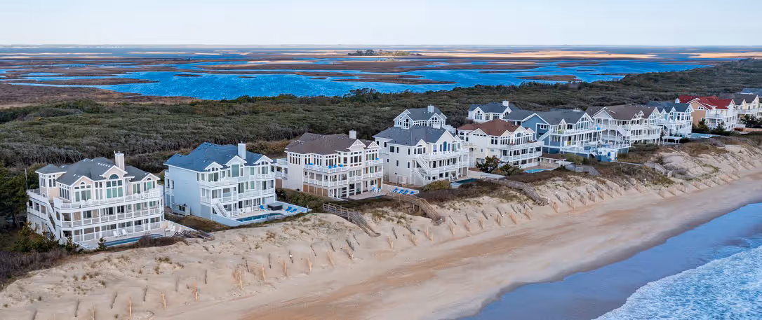 Aerial view of beachfront houses along sandy shore with ocean waves and marshland in the background.