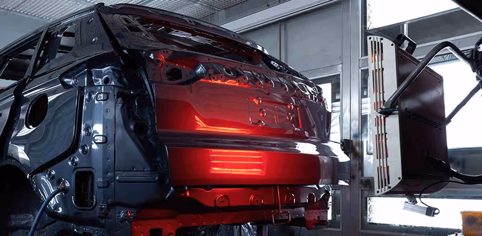 Partially assembled red car body on a production line inside a factory, with robotic equipment nearby.