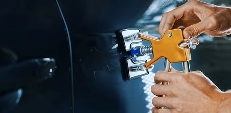 Hands using a yellow dent puller tool on a black car door to repair a dent.