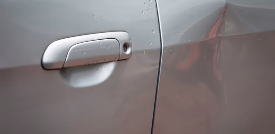 Close-up of a silver car door with a dent near the handle and another dent near the door edge.