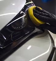 Gloved hand polishing a vehicle's headlight with a yellow foam applicator pad.