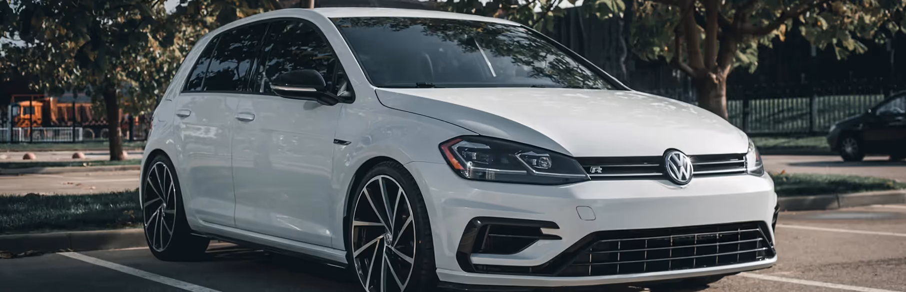 White Volkswagen Golf R parked in a shaded parking lot near trees.