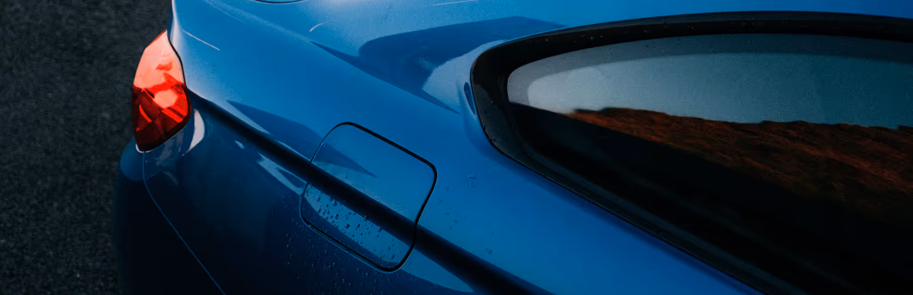 Close-up of a blue car's rear side showing the fuel door, tail light, and window reflection of a sunset.