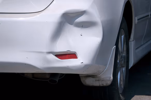 Close-up of the rear side of a dark-colored car with visible dents and scratches near the taillight and bumper.