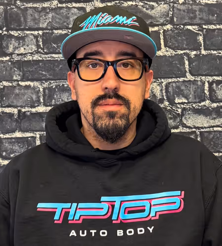 Man wearing black glasses, a Miami cap, and a black Tip Top Auto Body hoodie standing against a brick wall background.