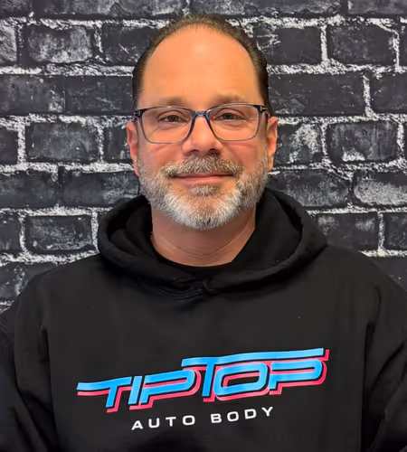 Man wearing glasses and a black hoodie with 'TIP TOP AUTO BODY' printed on it, standing against a dark brick wall.