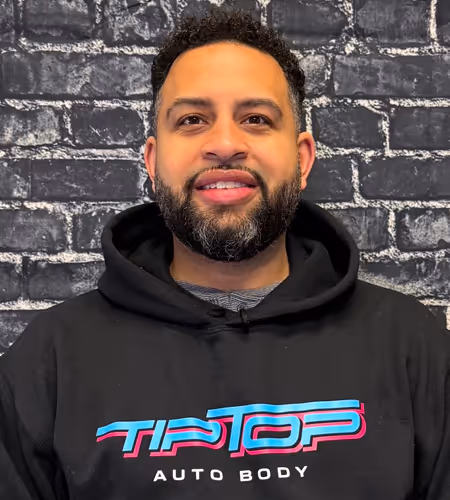 Smiling man with short curly hair and beard wearing a black Tip Top Auto Body hoodie against a dark brick wall background.