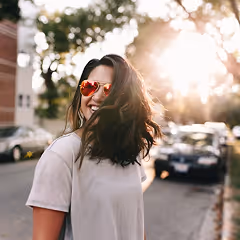 Smiling woman with long dark hair wearing red heart-shaped sunglasses and a white t-shirt outdoors at sunset.