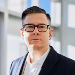 Man with dark hair and black glasses wearing a dark blazer and white shirt indoors.