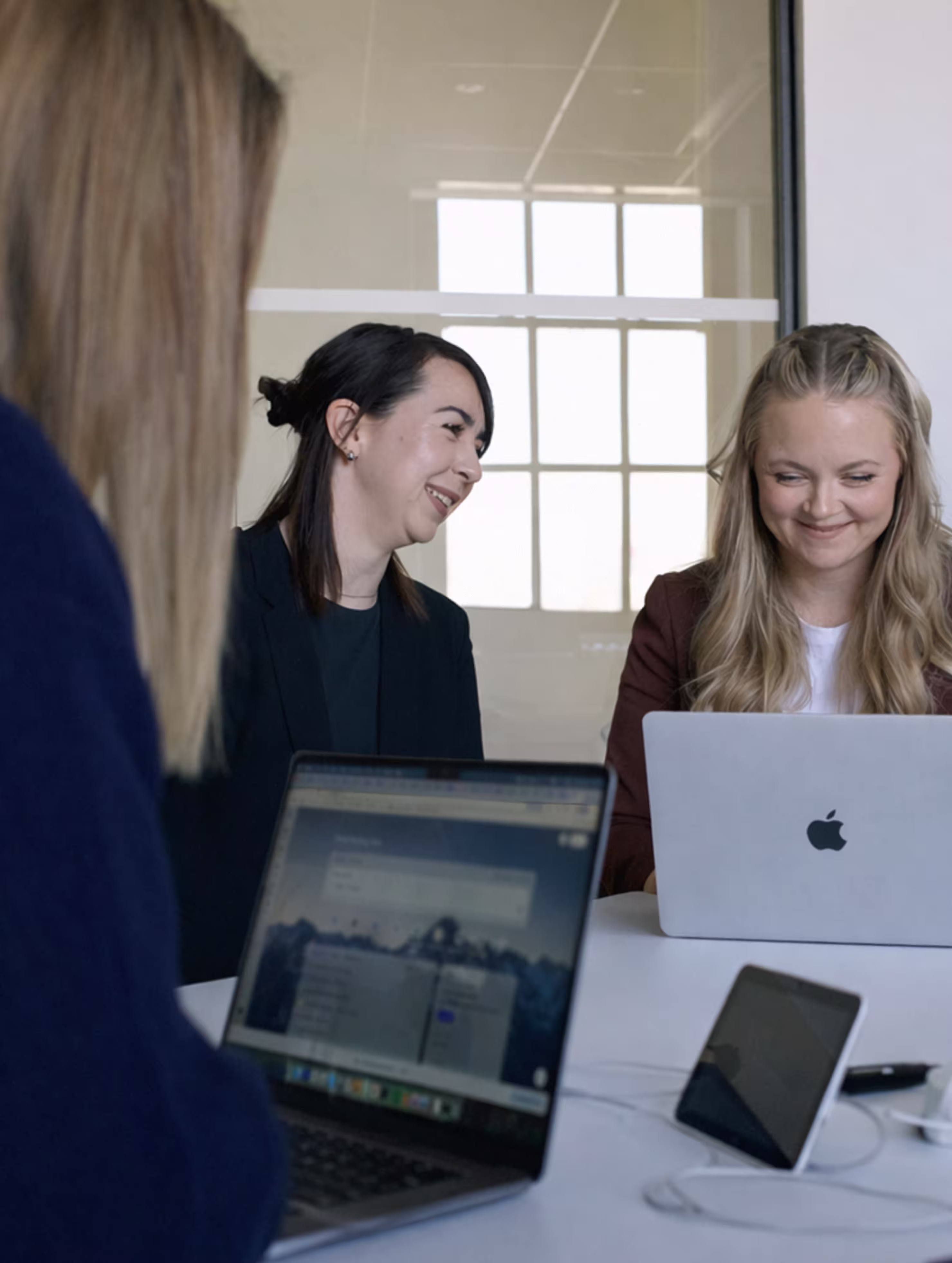 Three women sitting at a table with laptops, two smiling and engaged in conversation in a modern office.