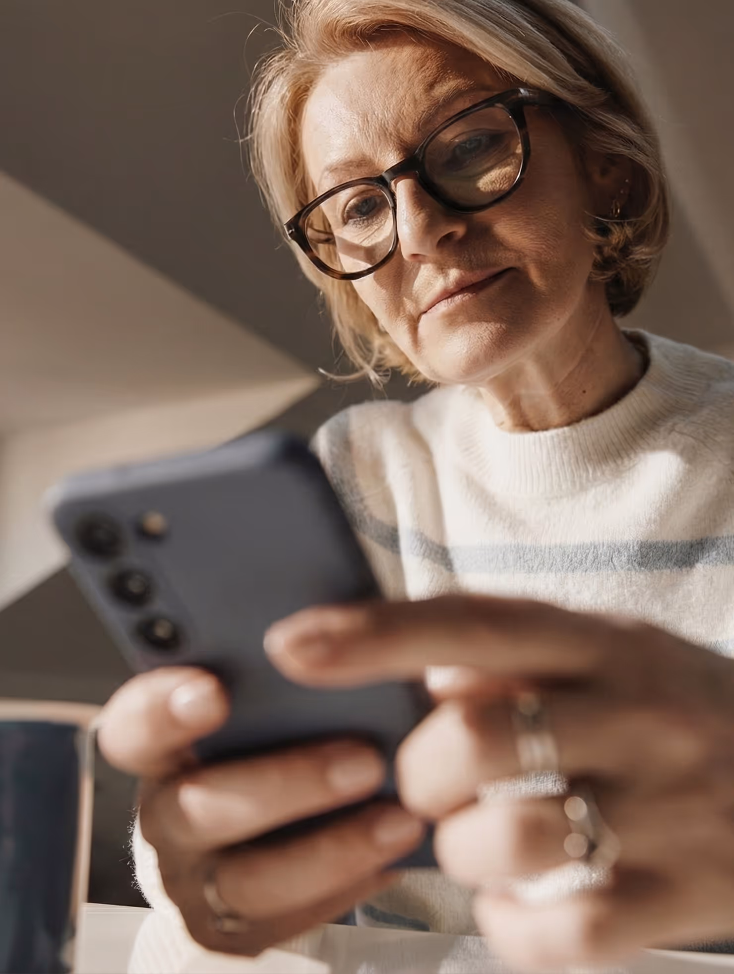 An older woman in glasses and a cream sweater focuses intently on her smartphone as she taps the screen, reviewing her medical bill, and going through the payment plan enrollment workflow.
