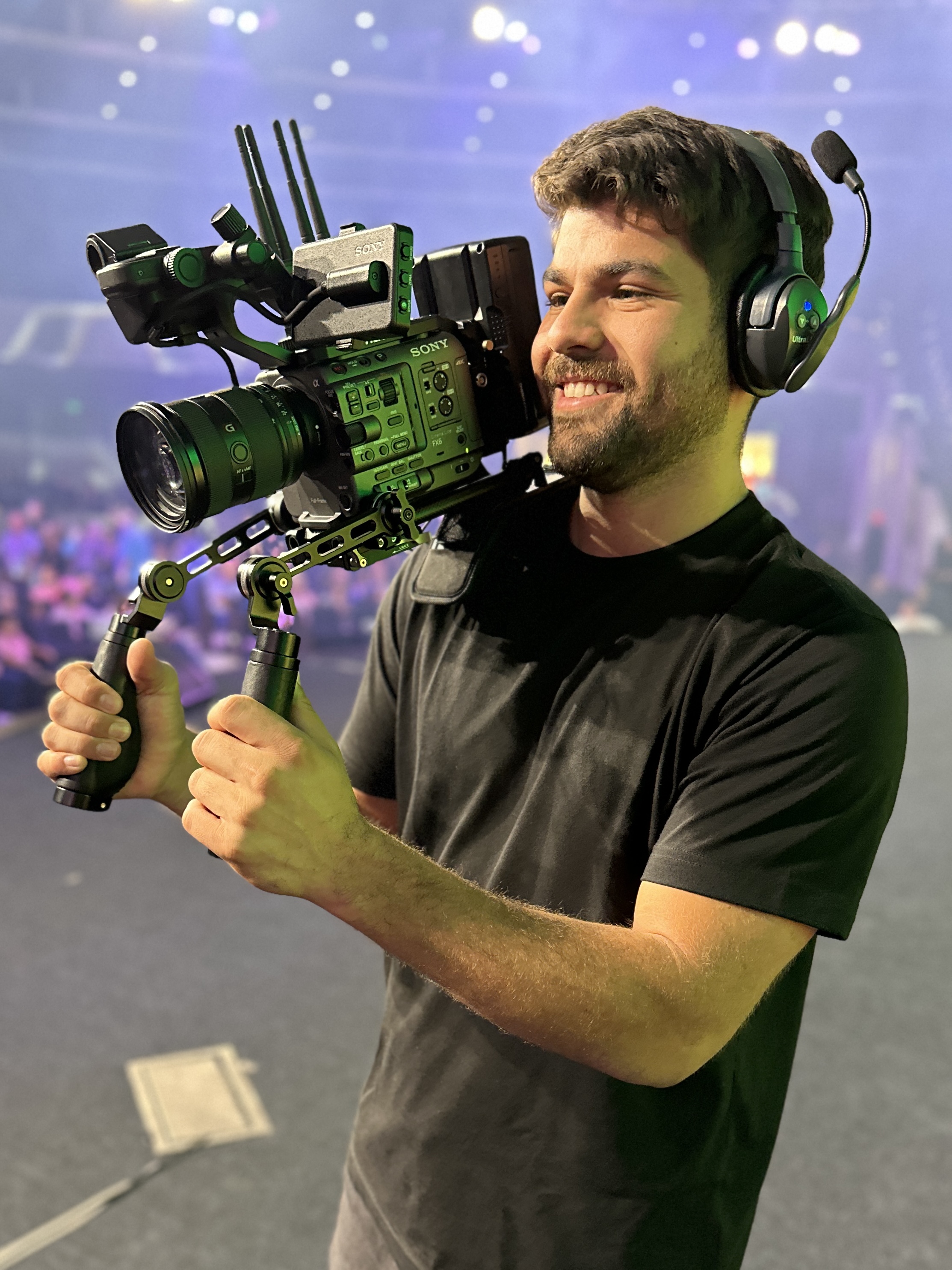 Smiling man wearing headphones and holding a professional Sony video camera rig on his shoulder in a blurred indoor venue.