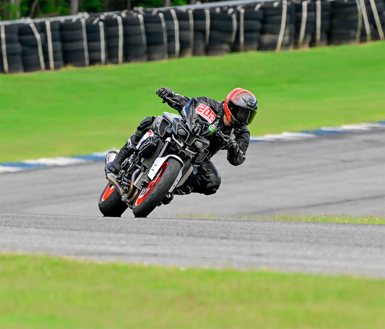 Motorcycle racer wearing black gear and an orange helmet leaning into a turn on a racetrack with grass and tire barriers in the background.