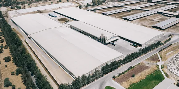 Vista aérea de una granja industrial con varios edificios grandes y caminos rodeados de árboles y áreas verdes.