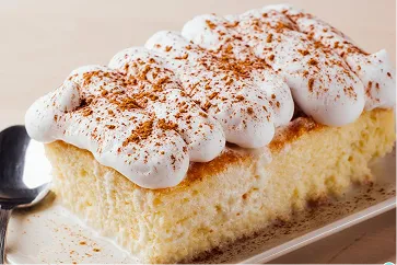 Rebanada de pastel tres leches con crema batida espolvoreada con canela.