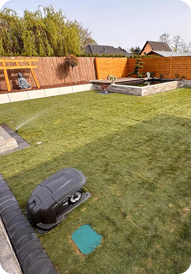 Garten mit grünem Rasen, automatischem Rasenmähroboter, springender Bewässerung und dekorativem Wasserbecken vor Holzzaun.