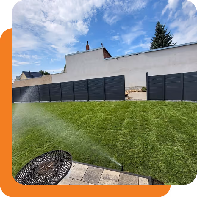 Ein gepflegter Rasen wird von einem automatischen Sprinkler bewässert, im Hintergrund sieht man eine moderne, hohe schwarze Zaunanlage und ein helles Gebäude unter blauem, bewölktem Himmel.