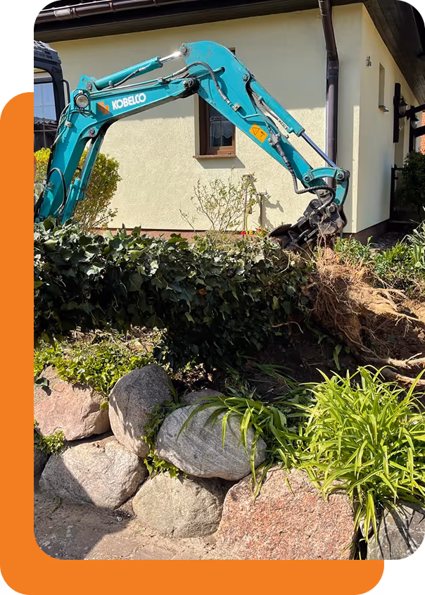 Grüner Kobelco-Bagger hebt eine große Wurzel aus einem bepflanzten Garten vor einem Haus mit gelber Fassade.