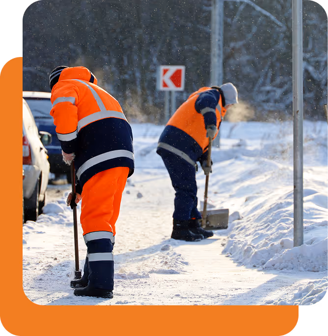 Zwei Arbeiter in orangefarbenen Warnkleidung räumen Schnee von einem Gehweg bei kaltem Wetter.