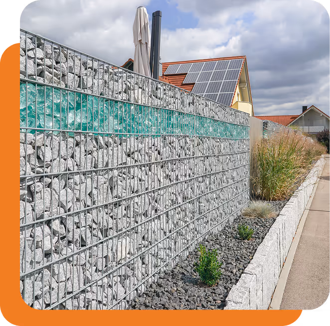 Gabionen-Mauer mit grauen Steinen und einem Streifen aus grünen Glassteinen vor einem Haus mit Solarpaneelen auf dem Dach.