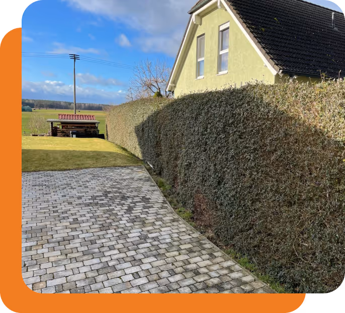 Gepflasterter Weg neben einem grünen Haus mit Hecke entlang des Weges und weitem Blick auf Felder unter blauem Himmel.