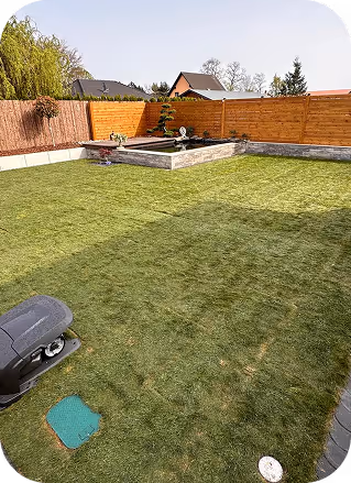 Ein gepflegter Garten mit grünem Rasen, einem Holzzaun und einem kleinen Hochbeet in einer sonnigen Ecke.