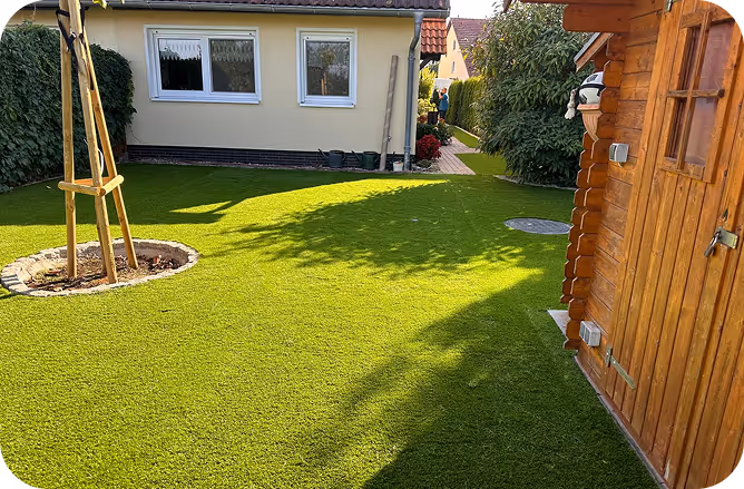 Grüner Rasen im Garten mit einem neu gepflanzten Baum und einem kleinen gelben Haus im Hintergrund.