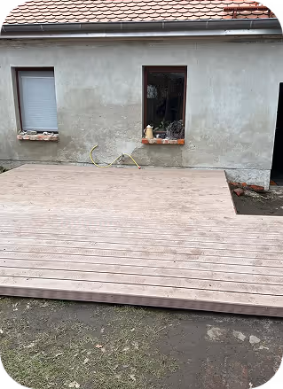 Hauswand mit zwei Fenstern und neu verlegtem Holzdeck davor, teilweise Erdreich sichtbar.