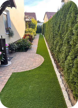 Enger Gartenweg mit Kopfsteinpflaster und gepflegtem Rasen neben hoher Hecke und bepflanztem Hausfassade.