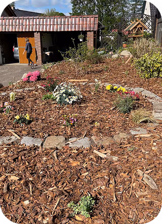 Gartenbeet mit frischen Blumen und Holzmulch vor einem Doppelgarage mit offener Tür, eine Person mit einem Kind geht hinein.