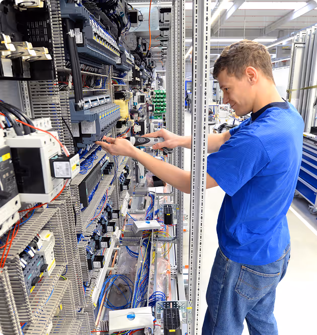 Ein Techniker in blauem T-Shirt arbeitet an einem Schaltschrank mit vielen Kabeln und elektronischen Komponenten in einer hellen Industrieumgebung.