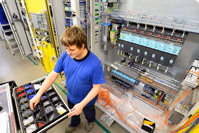 Techniker in blauem T-Shirt arbeitet an elektrischem Schaltschrank mit Werkzeug und Kabeln.