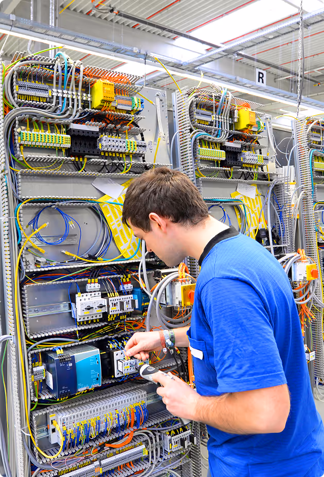 Ein Elektriker in blauem T-Shirt arbeitet an einem großen Schaltschrank mit vielen farbigen Kabeln und Steuergeräten.
