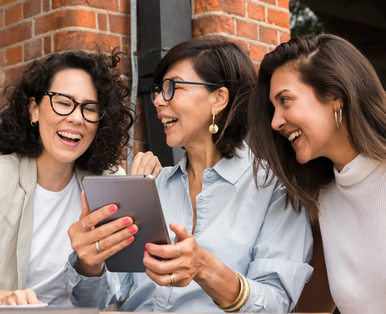 Three women smiling and looking at a tablet together while sitting by a brick wall.