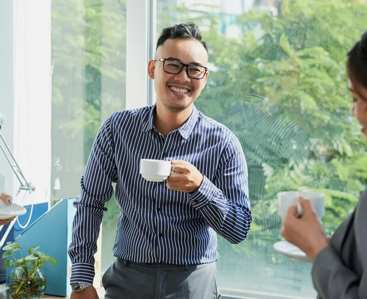 Smiling man with glasses in a striped shirt holding a white coffee cup standing near a window during a conversation.