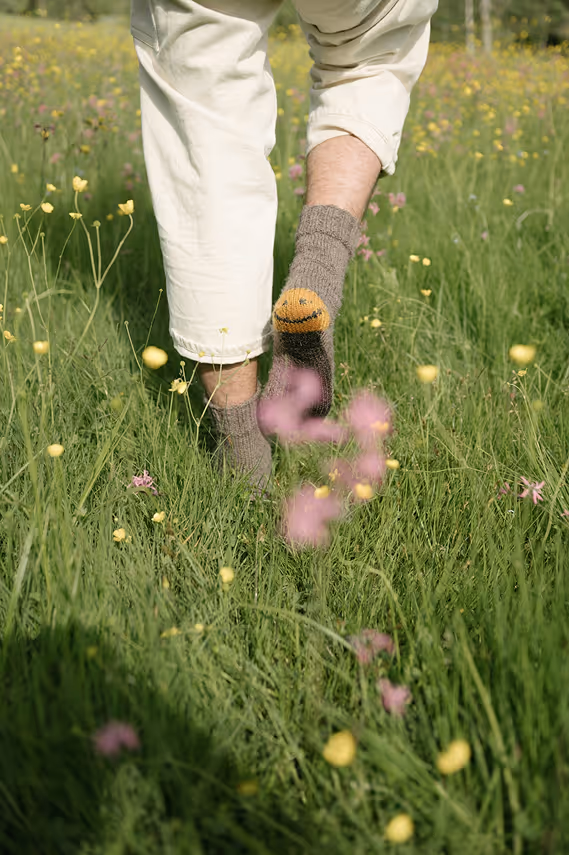 Person trägt braune Socken mit lachendem Gesicht, steht auf einer Wiese mit gelben und rosa Blumen.