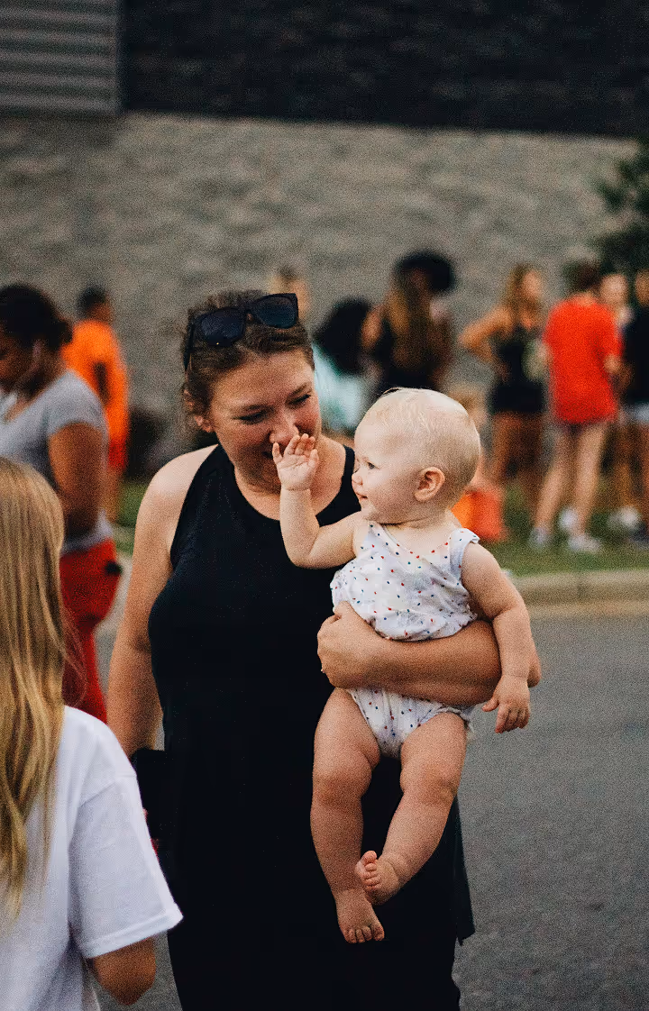 Frau hält ein lächelndes Baby in einer hellen Strampler und winkt einer Person mit langem Haar zu.